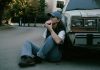 Woman savoring a donut while sitting against a vintage car in a serene street setting.