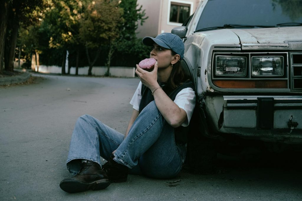 Woman savoring a donut while sitting against a vintage car in a serene street setting.