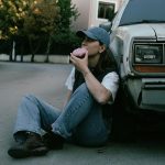 Woman savoring a donut while sitting against a vintage car in a serene street setting.