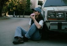Woman savoring a donut while sitting against a vintage car in a serene street setting.