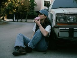 Woman savoring a donut while sitting against a vintage car in a serene street setting.