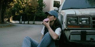 Woman savoring a donut while sitting against a vintage car in a serene street setting.