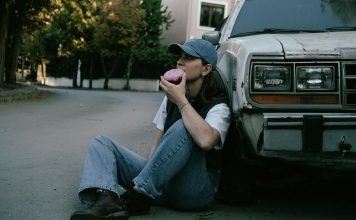 Woman savoring a donut while sitting against a vintage car in a serene street setting.