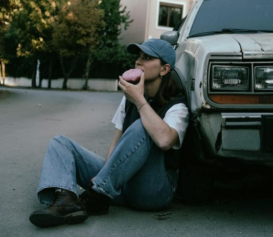 Woman savoring a donut while sitting against a vintage car in a serene street setting.