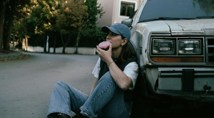 Woman savoring a donut while sitting against a vintage car in a serene street setting.