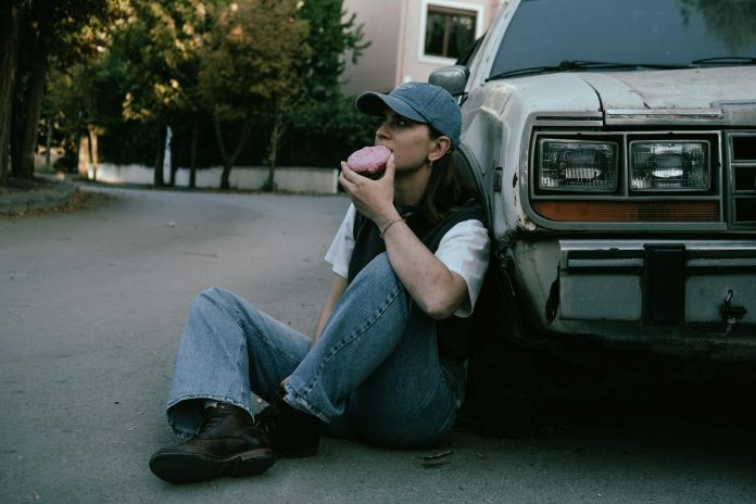 Woman savoring a donut while sitting against a vintage car in a serene street setting.