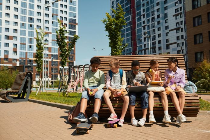 Children sitting on a bench, eating and sharing leisure time outdoors in a modern playground.