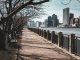 Bare trees line a waterfront walkway in New York City, with skyscrapers in the background.