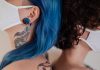Two women back to back wearing face masks, showcasing tattoos and colorful hair.