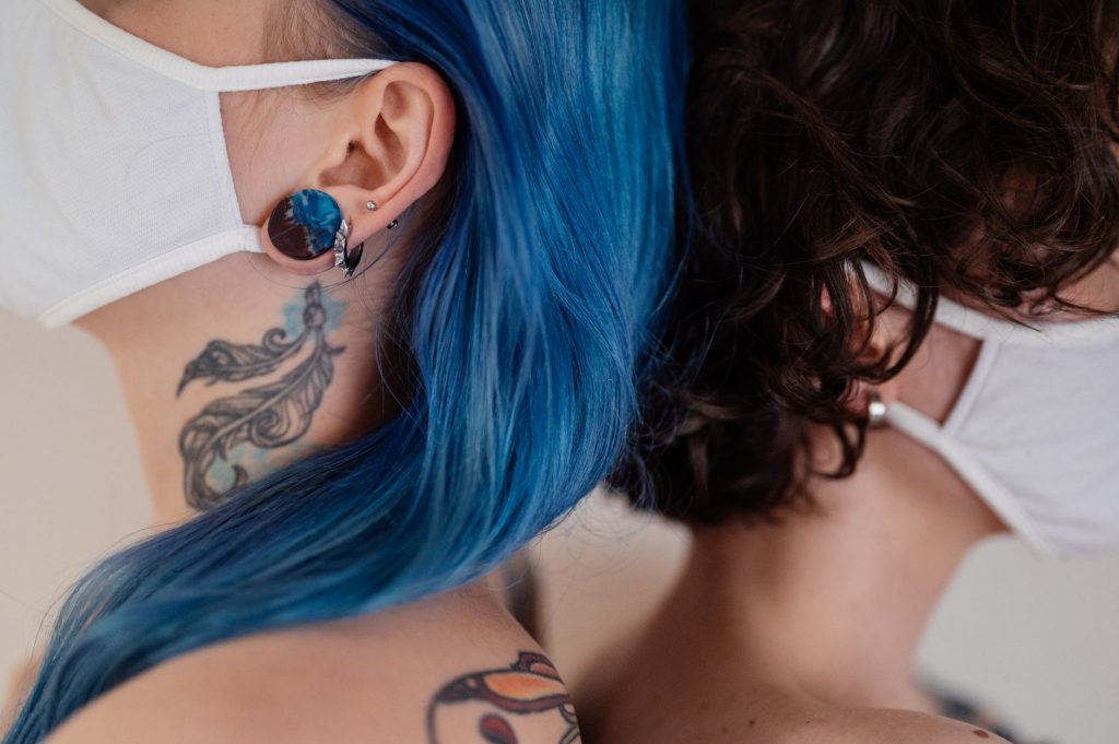 Two women back to back wearing face masks, showcasing tattoos and colorful hair.