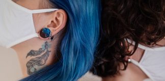 Two women back to back wearing face masks, showcasing tattoos and colorful hair.