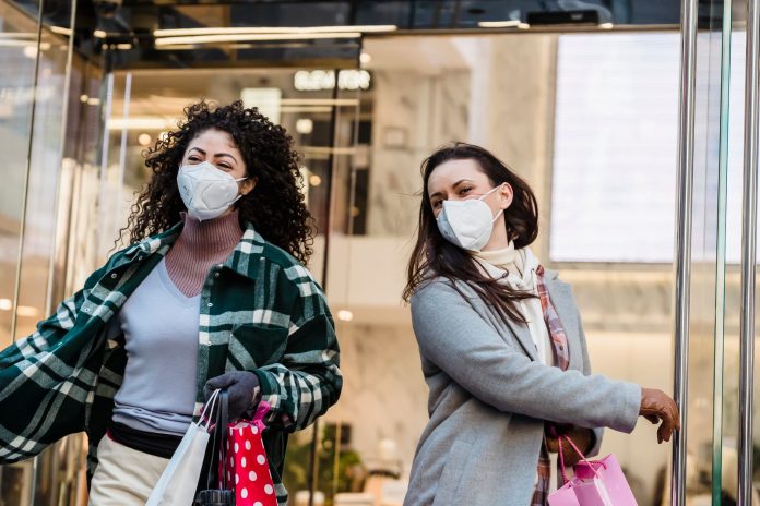 Low angle of woman with purchases in colorful shopping bags walking through doors of store during coronavirus pandemic