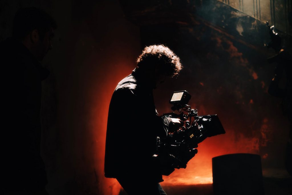 A professional cameraman captures footage in a creatively lit, moody setting, backlit by warm light.