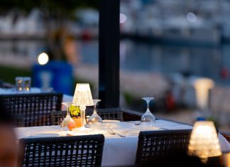 Charming outdoor restaurant setup with lamps and wine glasses by a marina at twilight.
