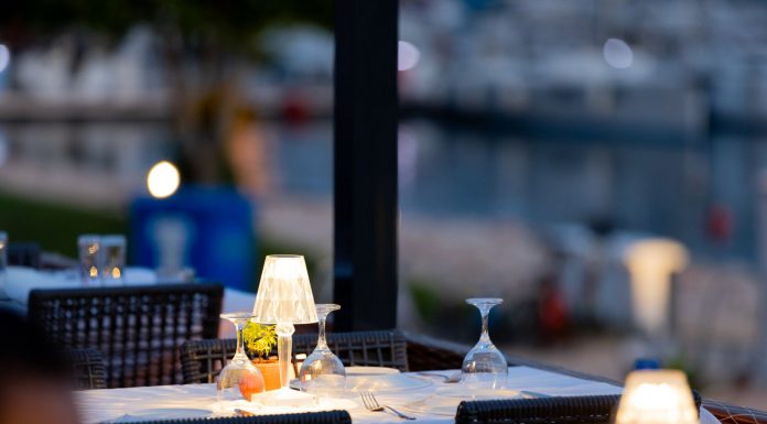 Charming outdoor restaurant setup with lamps and wine glasses by a marina at twilight.