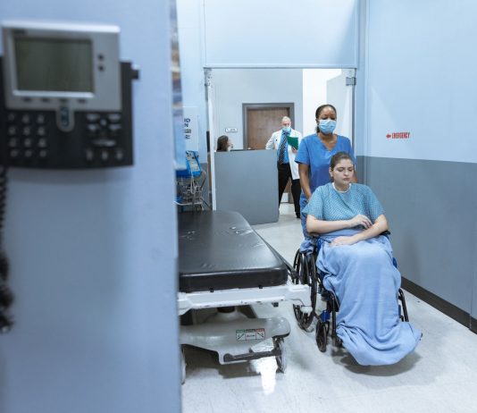 A nurse helps a patient in a wheelchair down a hospital corridor, reflecting care and medical professionalism.