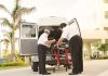 Two paramedics load medical equipment into a white ambulance outside a hospital in a tropical setting.