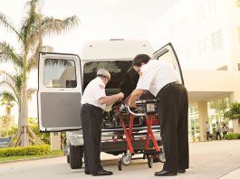 Two paramedics load medical equipment into a white ambulance outside a hospital in a tropical setting.