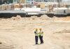 Two workers discussing plans on a sandy construction site, wearing safety gear.