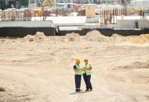 Two workers discussing plans on a sandy construction site, wearing safety gear.