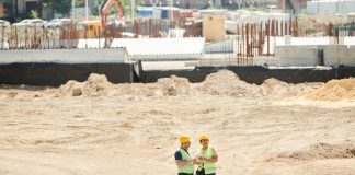 Two workers discussing plans on a sandy construction site, wearing safety gear.