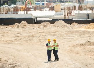 Two workers discussing plans on a sandy construction site, wearing safety gear.