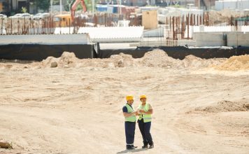 Two workers discussing plans on a sandy construction site, wearing safety gear.
