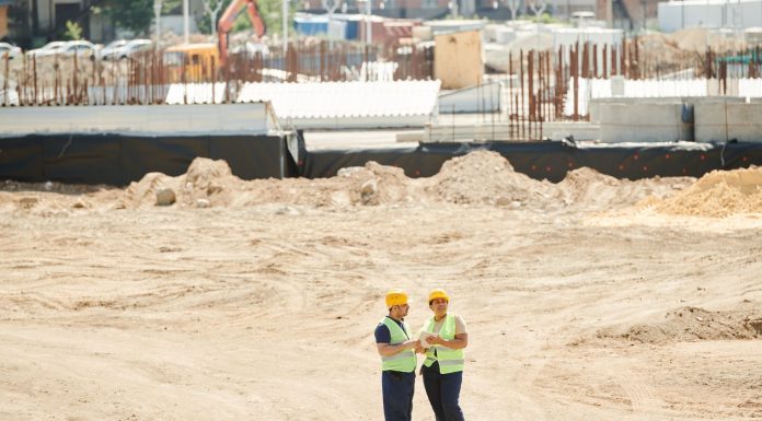 Two workers discussing plans on a sandy construction site, wearing safety gear.