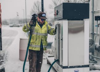 A man wearing a jacket refuels a truck at a gas station during winter with snow falling.