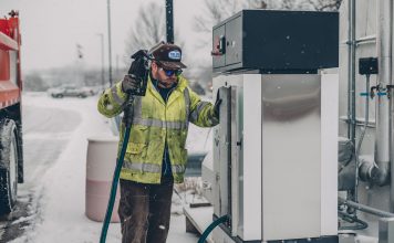 A man wearing a jacket refuels a truck at a gas station during winter with snow falling.