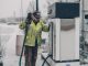 A man wearing a jacket refuels a truck at a gas station during winter with snow falling.