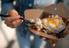 Close-up of a sunny side up egg in a takeout box being eaten with a fork.