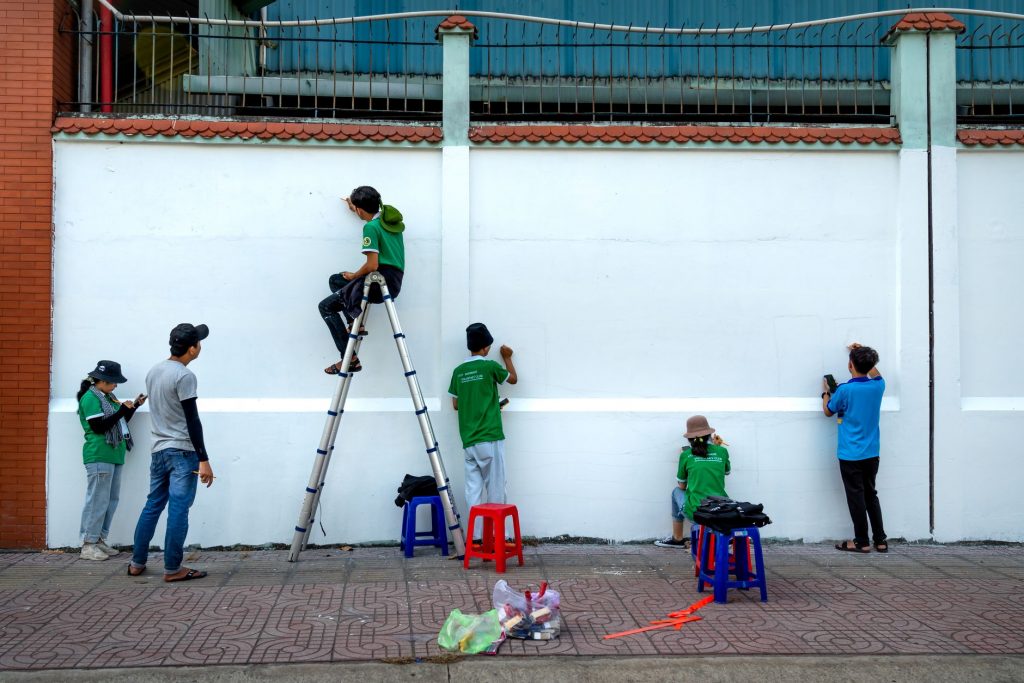 Group of artists painting a mural on a public wall outdoors using ladders and stools.