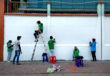 Group of artists painting a mural on a public wall outdoors using ladders and stools.