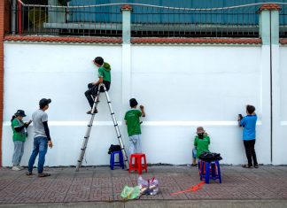 Group of artists painting a mural on a public wall outdoors using ladders and stools.