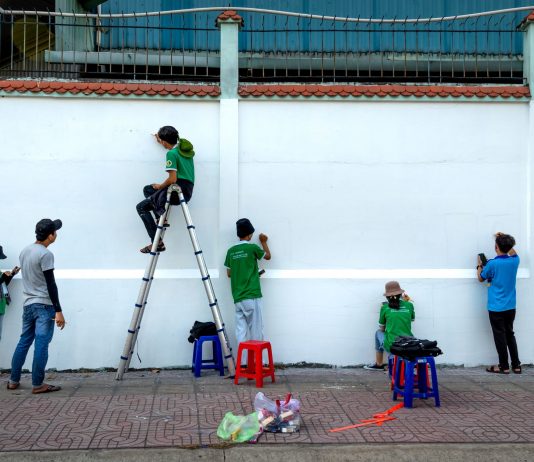 Group of artists painting a mural on a public wall outdoors using ladders and stools.