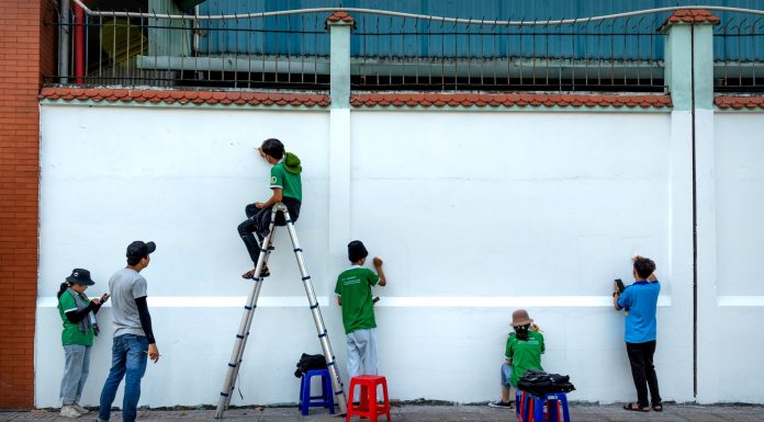 Group of artists painting a mural on a public wall outdoors using ladders and stools.