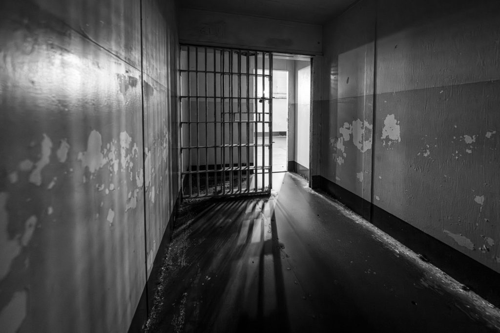 Moody black and white photo of a prison cell with an open barred door, evoking solitude.