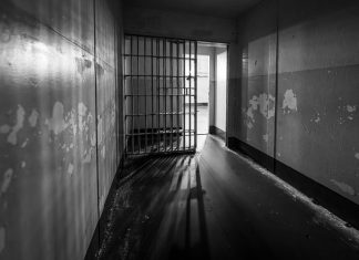 Moody black and white photo of a prison cell with an open barred door, evoking solitude.