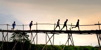 Unrecognizable active ethnic kids jogging on footbridge above lake while working out under sunset sky