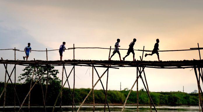 Unrecognizable active ethnic kids jogging on footbridge above lake while working out under sunset sky
