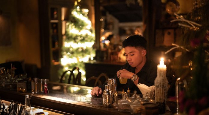 Hoboken’s The Waiting Room Hosts Xmas in July Event Young man enjoying a drink in a cozy bar with Christmas lights and decor.