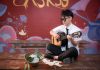 Young musician playing acoustic guitar indoors, busking next to a helmet with money.