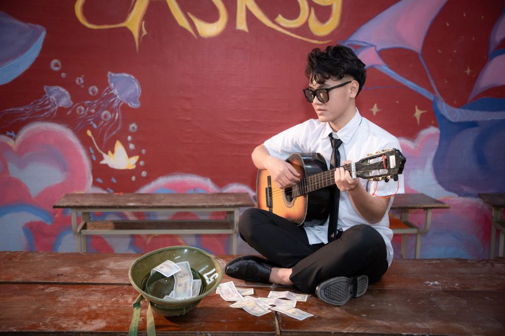 Young musician playing acoustic guitar indoors, busking next to a helmet with money.