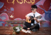 Young musician playing acoustic guitar indoors, busking next to a helmet with money.