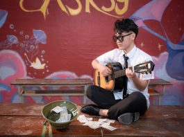 Young musician playing acoustic guitar indoors, busking next to a helmet with money.