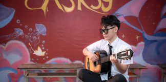 Young musician playing acoustic guitar indoors, busking next to a helmet with money.