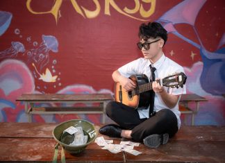Young musician playing acoustic guitar indoors, busking next to a helmet with money.