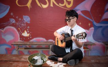 Young musician playing acoustic guitar indoors, busking next to a helmet with money.