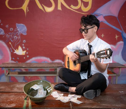Young musician playing acoustic guitar indoors, busking next to a helmet with money.